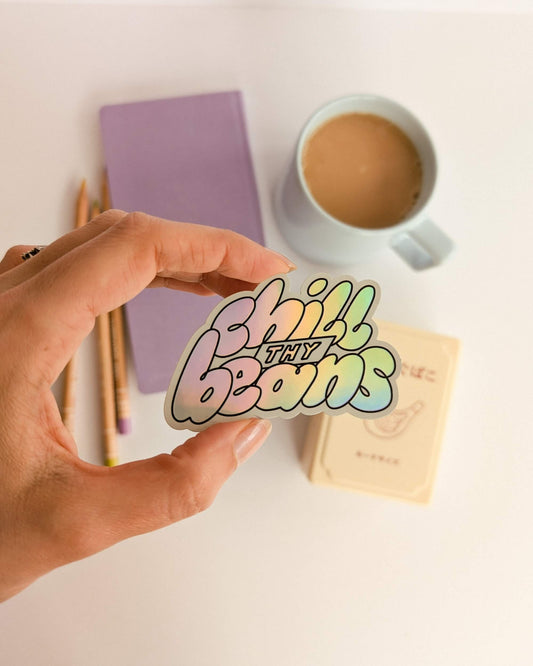Hand holding a 'Chill Thy Beans' sticker with a coffee cup and notebook in the background.