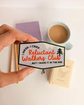 Hand holding a sticker with 'Reluctant Walkers Club' text, coffee cup in background