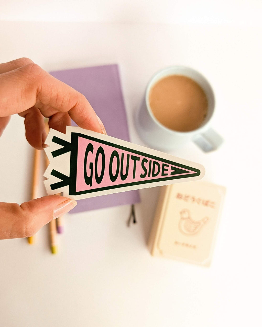 Hand holding a 'GO OUTSIDE' sticker with a cup of coffee and stationery in the background