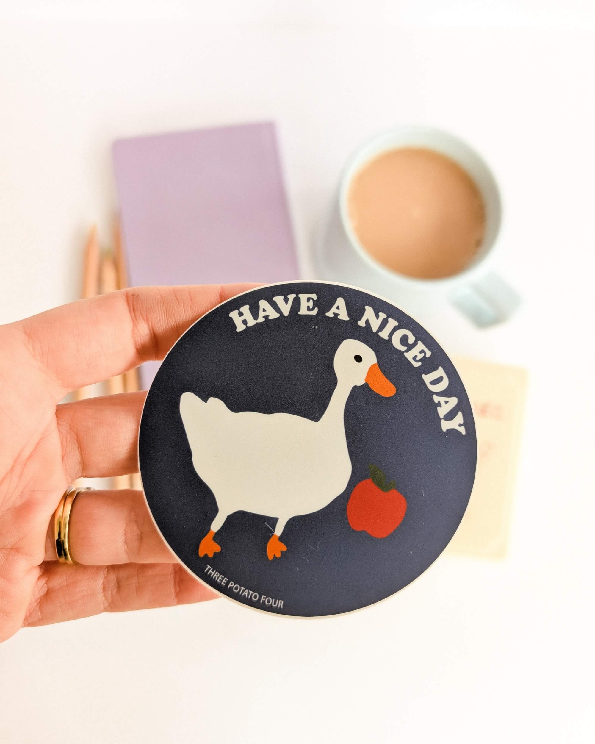 Sticker with a duck design and 'Have a nice day' text held by a hand, with a cup of coffee and notebook in the background.