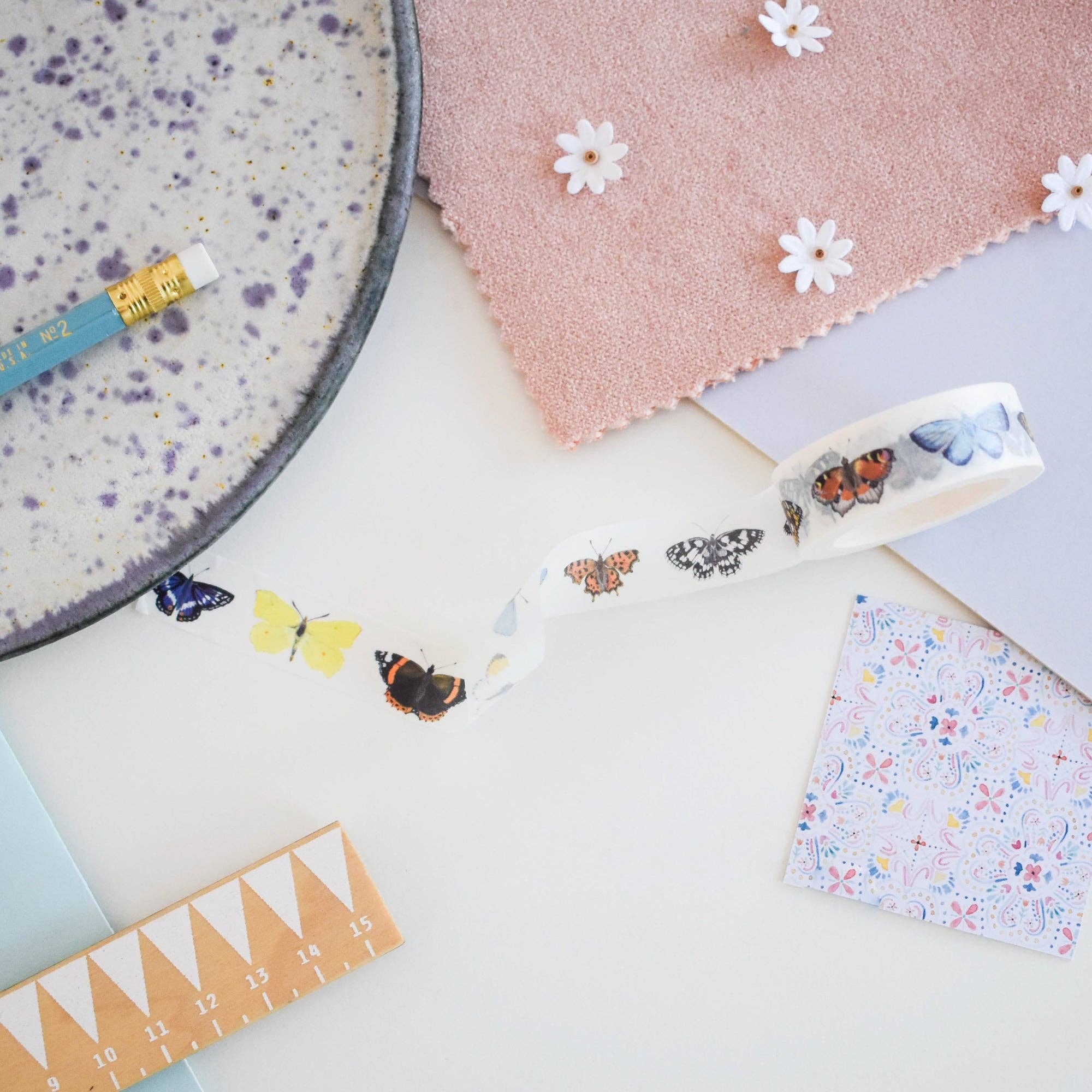 Stationery items including a pencil, washi tape with butterfly designs, and floral paper on a light surface.
