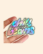 Holographic sticker with 'Chill Thy Beans' text held in a hand against a beige background