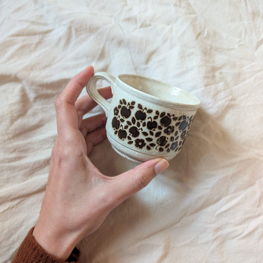 Hand holding a ceramic cup with black floral patterns on a soft, light-colored fabric background